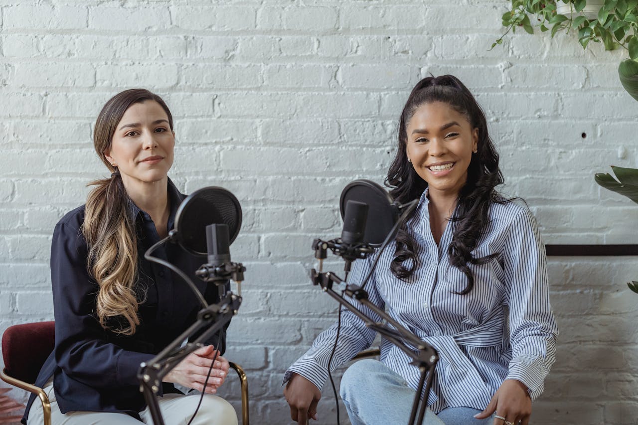 Two diverse women smiling in podcast studio with microphones.