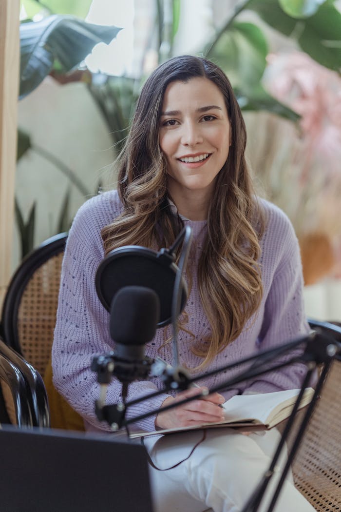 Cheerful female radio host looking at camera while sitting in chair with opened notebook near professional microphone in light room